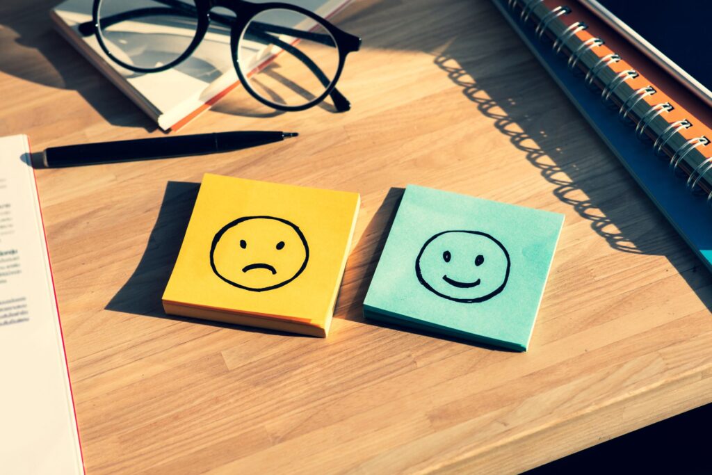 Dwie karteczki samoprzylepne na biurku: jedna żółta ze smutną buźką, druga niebieska z uśmiechniętą buźką; obok okulary, długopis i zeszyt