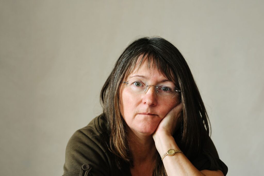 Kobieta w średnim wieku w okularach, opierająca głowę na dłoni, patrzy zamyślonym wzrokiem w stronę obiektywu.