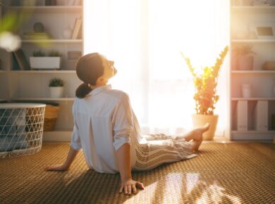 Kobieta siedzi w pokoju na przeciwko okna, przez które wpada słońce. Odpoczywa.