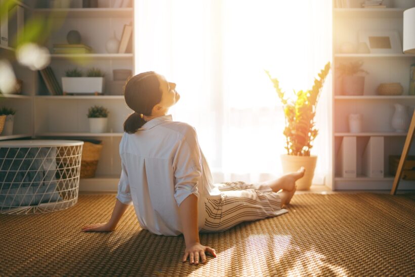 Kobieta siedzi w pokoju na przeciwko okna, przez które wpada słońce. Odpoczywa.