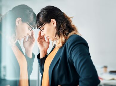 Młoda kobieta w okularach, oparta czołem o szybę, z zamkniętymi oczami i ręką na czole – wygląda na zestresowaną lub zamyśloną.