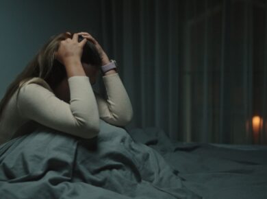 Kobieta siedzi na łóżku i trzyma się za głowę. Jest noc. Ciemno.