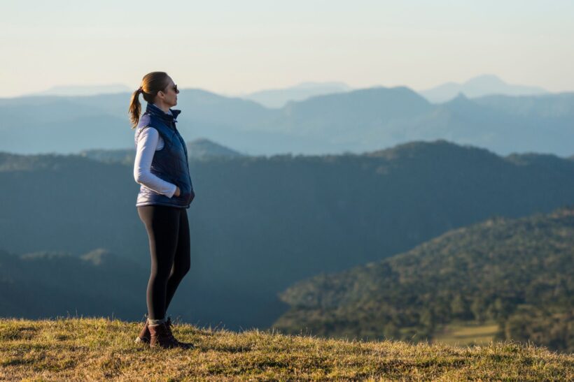 Kobieta w sportowym stroju stojąca na górskiej łące z rękami w kieszeniach, spoglądająca w dal na górskie pasma przy porannym świetle.