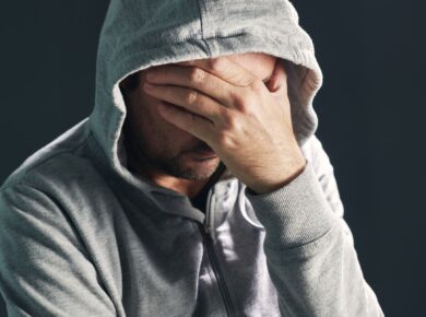 Mężczyzna w szarej bluzie z kapturem zasłania twarz dłonią, wyrażając smutek lub przygnębienie