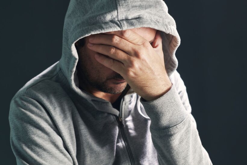Mężczyzna w szarej bluzie z kapturem zasłania twarz dłonią, wyrażając smutek lub przygnębienie