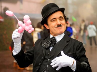 Artysta uliczny przebrany za Charliego Chaplina z różowym balonem w ręku, uśmiechnięty i puszczający oko, w tle rozmyta scena miejska