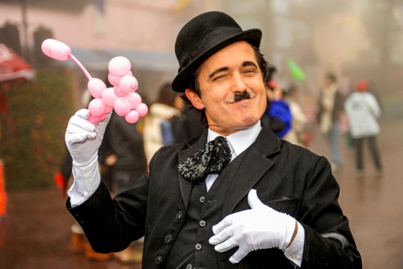 Artysta uliczny przebrany za Charliego Chaplina z różowym balonem w ręku, uśmiechnięty i puszczający oko, w tle rozmyta scena miejska