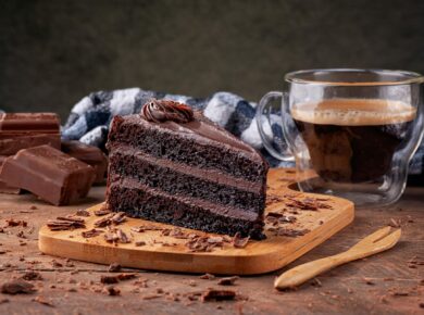 Kawałek ciemnego czekoladowego tortu z kremem na drewnianej desce, obok filiżanka kawy i kostki czekolady, w tle koc w kratę.