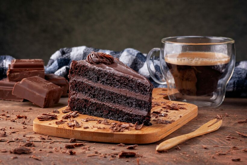 Kawałek ciemnego czekoladowego tortu z kremem na drewnianej desce, obok filiżanka kawy i kostki czekolady, w tle koc w kratę.