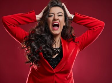 Kobieta w czerwonym garniturze z rozpuszczonymi włosami, trzymająca głowę w dłoniach i krzycząca z emocjami.