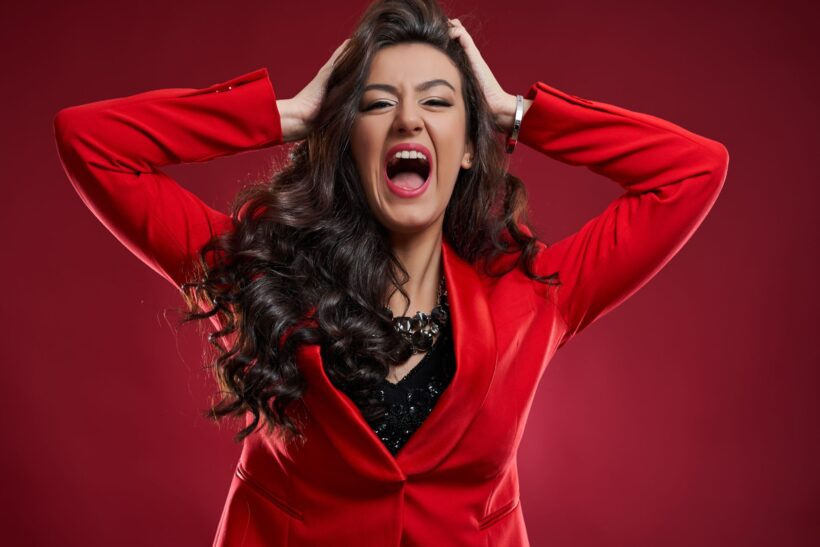 Kobieta w czerwonym garniturze z rozpuszczonymi włosami, trzymająca głowę w dłoniach i krzycząca z emocjami.