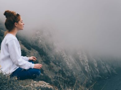 Kobieta siedząca w pozycji medytacyjnej na skalistym zboczu, otoczona mgłą, wpatrzona spokojnie w przestrzeń.
