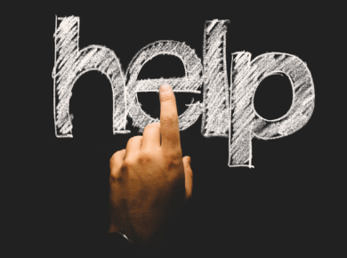 Dłoń dotykająca litery "e" w słowie "help" napisanym kredą na czarnej tablicy.