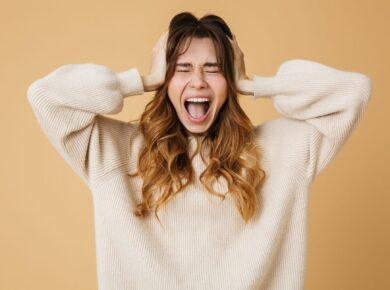 Kobieta w beżowym swetrze trzymająca się za głowę i krzycząca z zamkniętymi oczami, wyrażająca silne emocje, takie jak frustracja, złość lub stres. Tło w kolorze jasnego beżu.