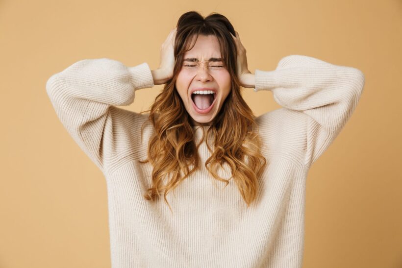 Kobieta w beżowym swetrze trzymająca się za głowę i krzycząca z zamkniętymi oczami, wyrażająca silne emocje, takie jak frustracja, złość lub stres. Tło w kolorze jasnego beżu.