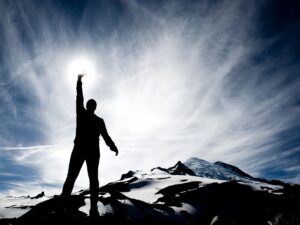 Sylwetka człowieka na szczycie góry sięgającego dłonią po słońce – symbol siły i odwagi