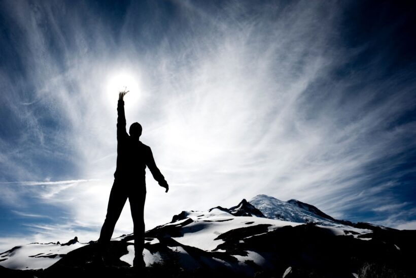 Sylwetka człowieka na szczycie góry sięgającego dłonią po słońce – symbol siły i odwagi