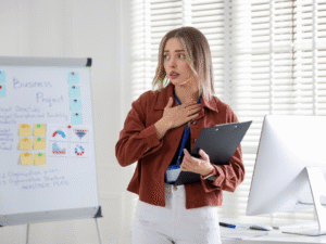Młoda kobieta stojąca przy tablicy projektowej w biurze trzyma dłoń na piersi, wyrażając strach i zdenerwowanie podczas wystąpienia.