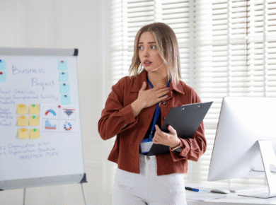 Młoda kobieta stojąca przy tablicy projektowej w biurze trzyma dłoń na piersi, wyrażając strach i zdenerwowanie podczas wystąpienia.