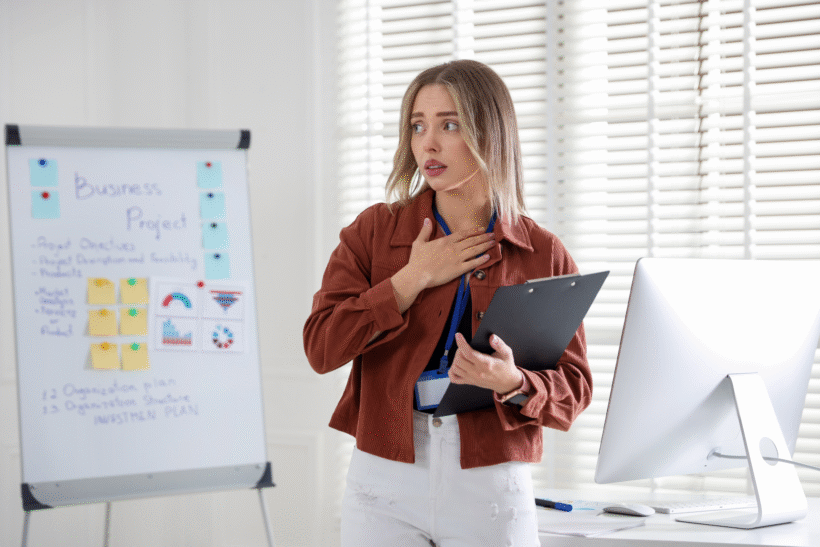 Młoda kobieta stojąca przy tablicy projektowej w biurze trzyma dłoń na piersi, wyrażając strach i zdenerwowanie podczas wystąpienia.