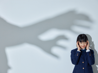 Dziewczyna w szkolnym mundurku zasłania uszy, a na ścianie widać cień ogromnych dłoni symbolizujących strach.
