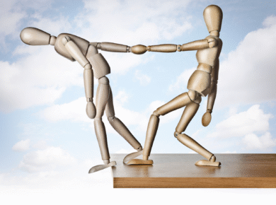 Drewniane figurki – jedna pomaga drugiej, chroniąc przed upadkiem – symbol wsparcia i relacji.