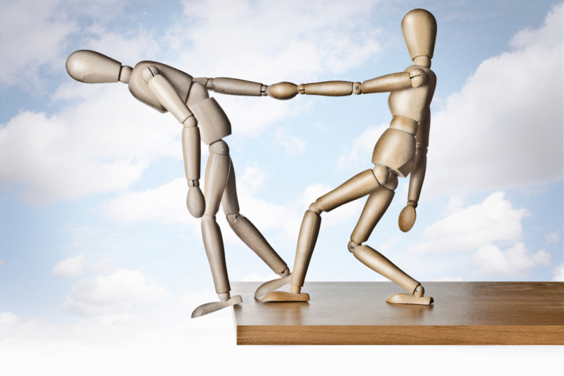 Drewniane figurki – jedna pomaga drugiej, chroniąc przed upadkiem – symbol wsparcia i relacji.