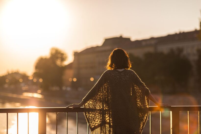 Kobieta oparta o barierkę mostu, patrząca w stronę zachodzącego słońca – obraz refleksji, spokoju i momentu, w którym samotność staje się przestrzenią do odzyskania siebie.